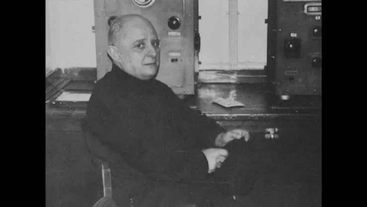 Ο Νίκος Καββαδίας κάθεται με σκούρο πουλόβερ ανάμεσα σε ηλεκτρονικές συσκευές ασυρμάτου