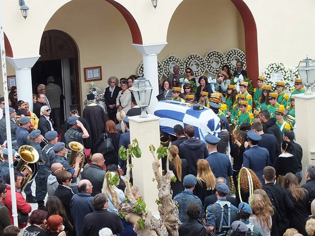 Πλήθος και στεφάνια έξω από την εκκλησία, με το φέρετρο της Άννας Πολλάτου καλυμμένο με ελληνική σημαία και μπάντα με πράσινες στολές