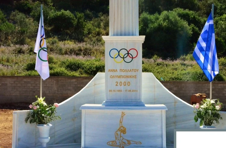 Το μαρμάρινο μνημείο της Άννας Πολλάτου με τους ολυμπιακούς κύκλους, την επιγραφή «Άννα Πολλάτου, Ολυμπιονίκης 2000» και δύο σημαίες