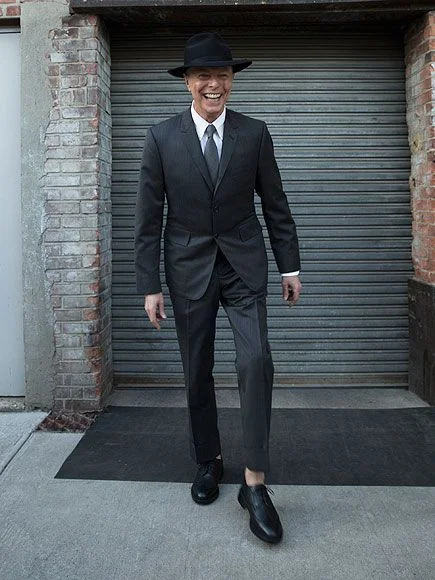 Ο David Bowie χαμογελαστός με σκούρο κοστούμι και καπέλο μπροστά σε μεταλλικό ρολό