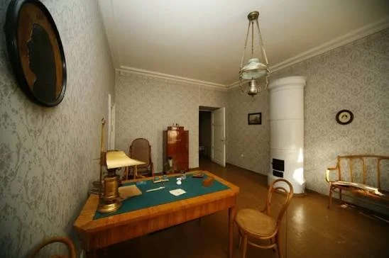 Δωμάτιο με γραφείο, καρέκλα, καναπέ και ψηλή λευκή κεραμική σόμπα.