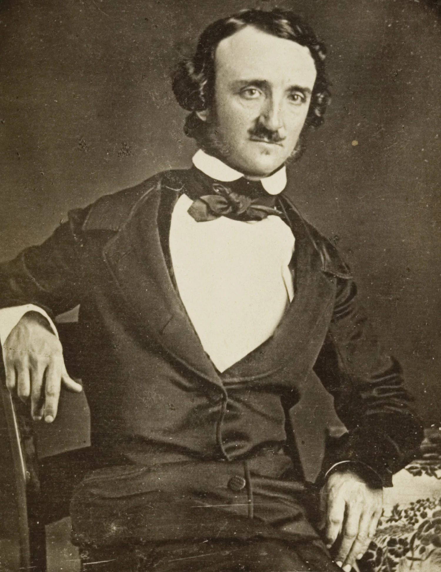 Ασπρόμαυρο πορτρέτο του Edgar Allan Poe καθιστού, με σκούρο σακάκι, λευκό πουκάμισο και παπιγιόν, με το χέρι ακουμπισμένο στο μπράτσο της καρέκλας