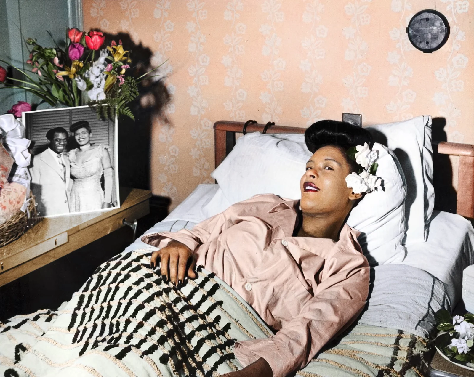 Η Billie Holiday ξαπλωμένη σε κρεβάτι, με λευκά λουλούδια στα μαλλιά και μια φωτογραφία στο κομοδίνο, σε χρωματισμένη φωτογραφία της δεκαετίας του 1940