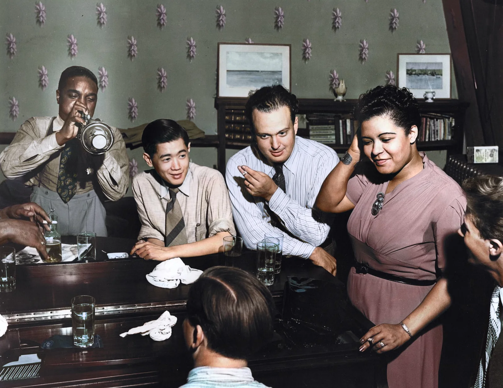 Η Billie Holiday και μουσικοί γύρω από ένα τραπέζι, ενώ ένας τρομπετίστας παίζει, σε χρωματισμένη φωτογραφία του 1939