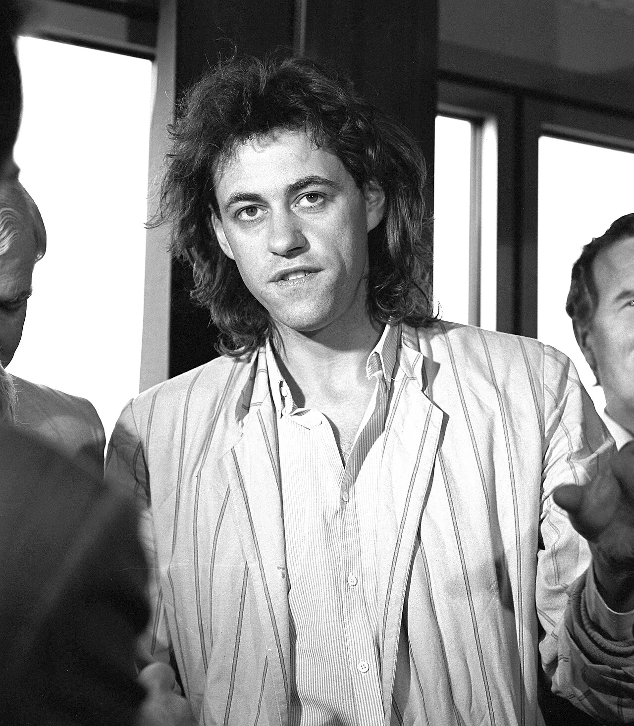 Ασπρόμαυρο πορτρέτο του Bob Geldof με ριγέ σακάκι, να μιλά μπροστά σε παράθυρα