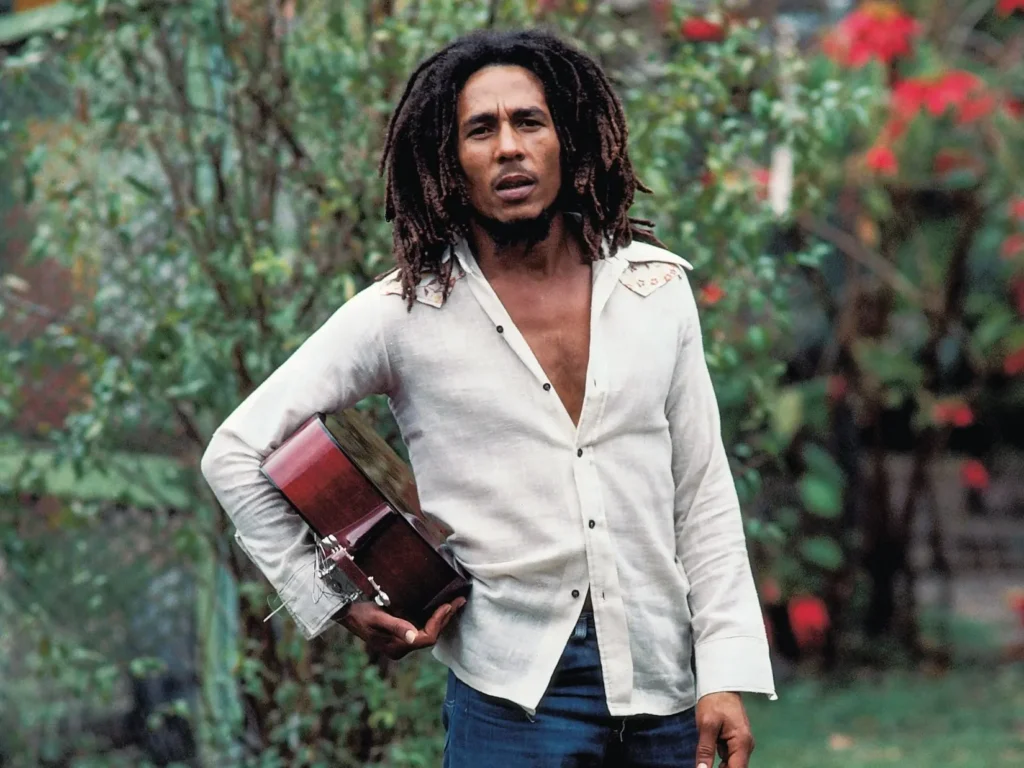 Έγχρωμο πορτρέτο του Bob Marley σε εξωτερικό χώρο με βλάστηση. Ο μουσικός φορά λευκό πουκάμισο και τζιν και κρατά ακουστική κιθάρα. Δεν προστίθενται συγκεκριμένο έτος, τόπος ή φωτογράφος, επειδή δεν επαληθεύτηκαν για αυτό το αρχείο.