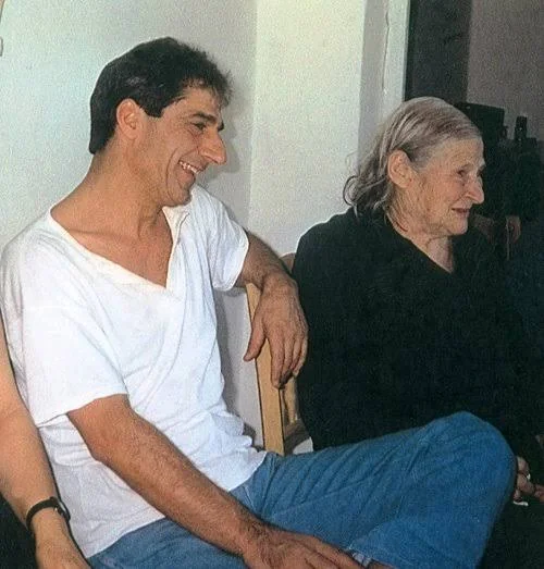 Άνδρας με λευκή μπλούζα και τζιν χαμογελά, καθισμένος δίπλα σε ηλικιωμένη γυναίκα με μαύρα ρούχα
