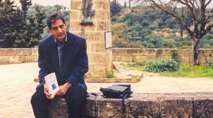 Ο Λιαντίνης καθισμένος σε πέτρινο χαμηλό τοίχο με ένα λευκό βιβλίο στα χέρια, μπροστά από πέτρινο μνημείο με επιγραφή και δέντρα πίσω του