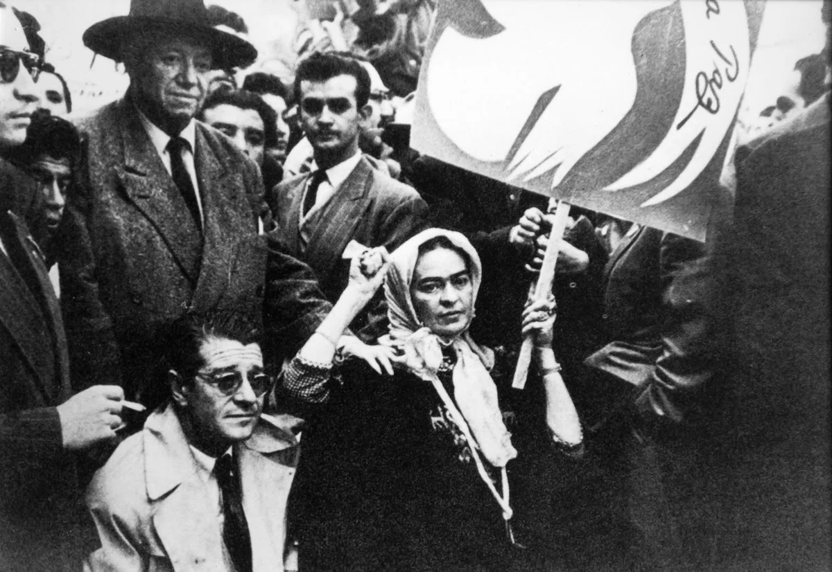Ασπρόμαυρη φωτογραφία από διαδήλωση: γυναίκα με μαντίλα στο κεφάλι ανασηκώνει τη γροθιά της μπροστά από πινακίδα, με όρθιο άνδρα με καπέλο πίσω της
