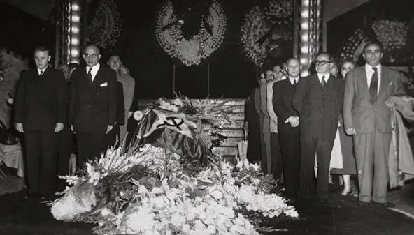 Ασπρόμαυρη φωτογραφία με φέρετρο καλυμμένο με σημαία με σφυροδρέπανο ανάμεσα σε λουλούδια και όρθιους άνδρες με κοστούμια