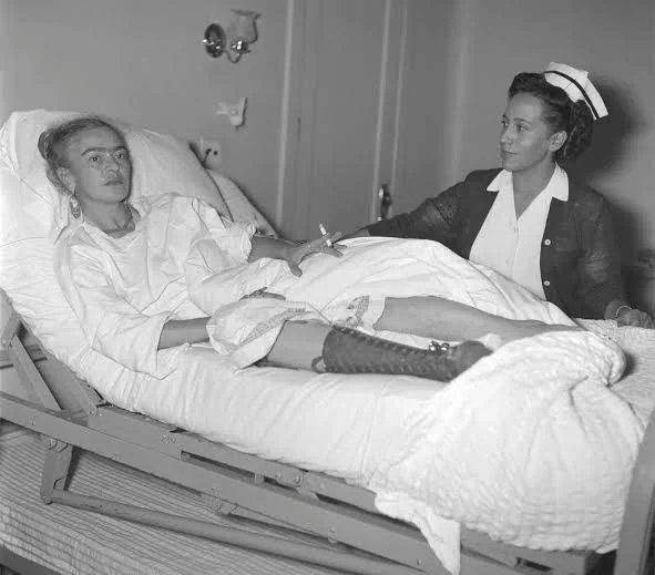Ασπρόμαυρη φωτογραφία της Frida Kahlo σε νοσοκομειακό κρεβάτι με λευκή ρόμπα και δεμένη μπότα στο πόδι, με νοσοκόμα δίπλα της