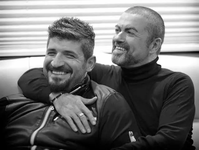 Ο George Michael αγκαλιάζει τον Fadi Fawaz ενώ οι δύο άνδρες χαμογελούν καθισμένοι σε καναπέ.