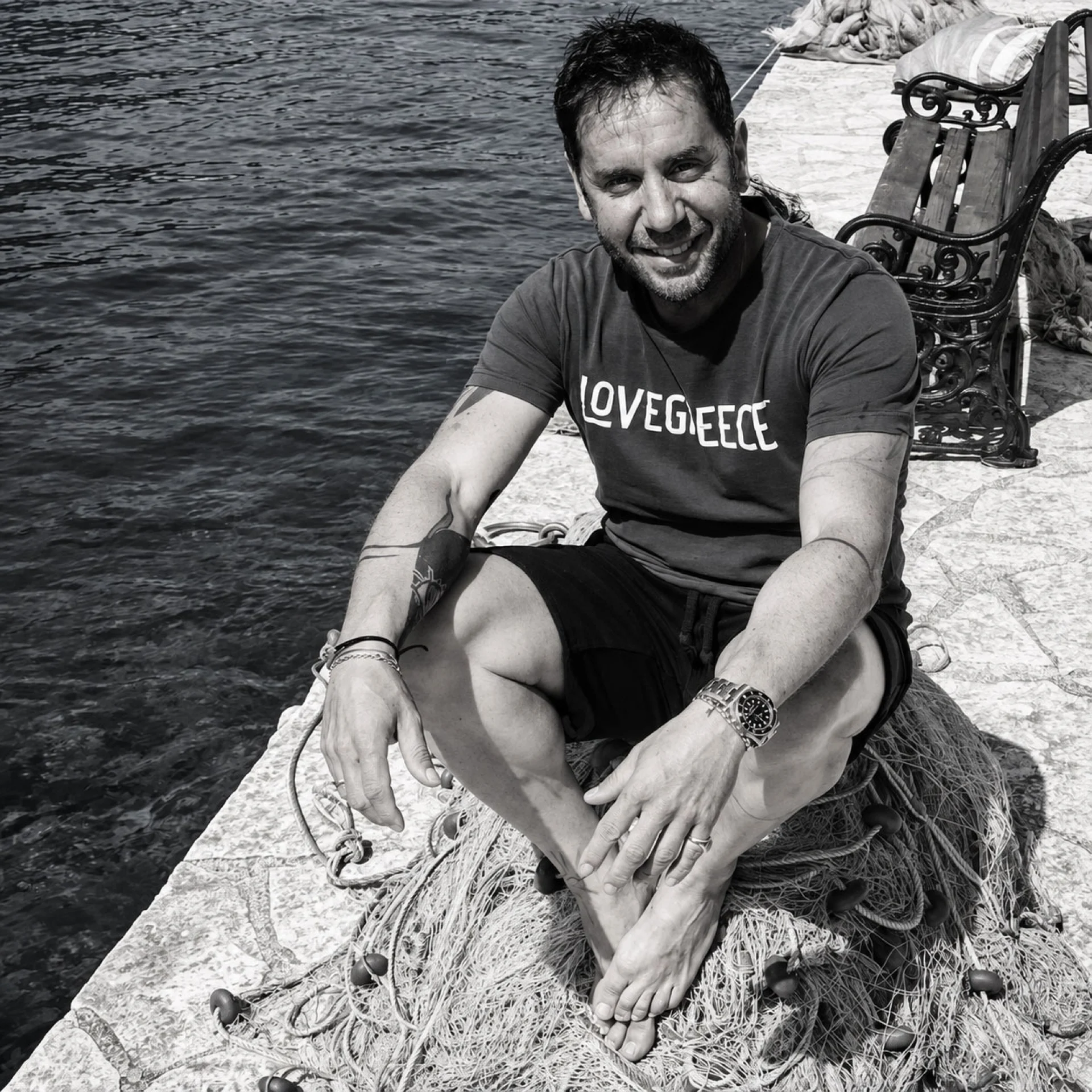 Ασπρόμαυρη φωτογραφία του Γιώργου Μαζωνάκη να κάθεται σταυροπόδι πάνω σε δίχτυα ψαρέματος στο λιμάνι, χαμογελώντας πλατιά