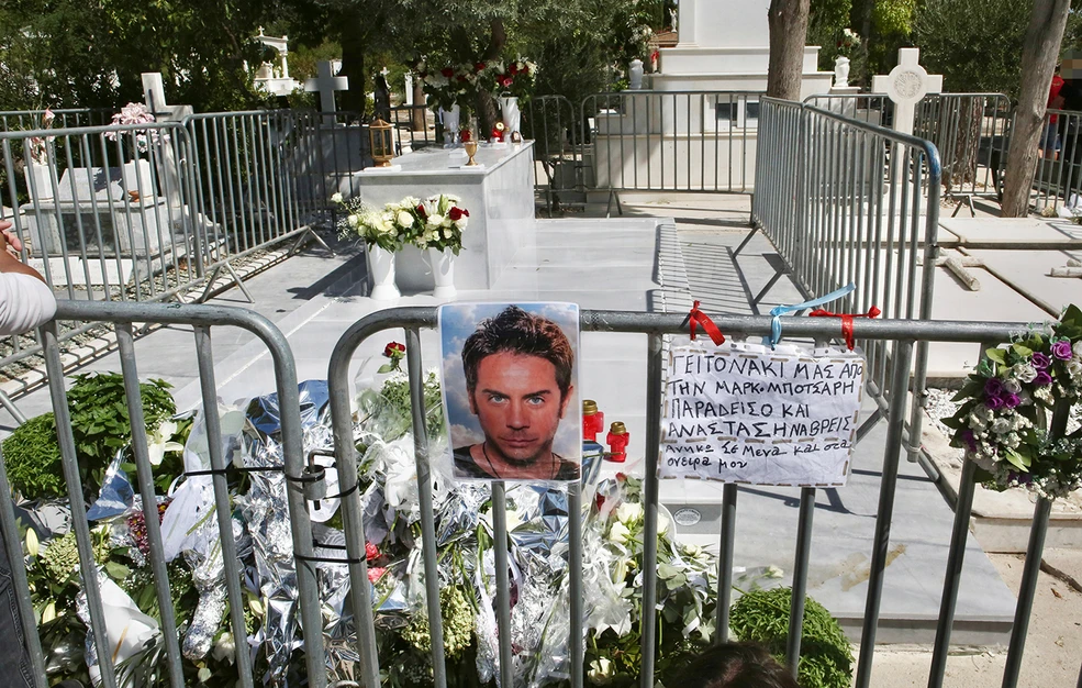 Ο τάφος του Γιώργου Μαζωνάκη γεμάτος λευκά λουλούδια. Στα κάγκελα είναι κρεμασμένη μια φωτογραφία του και ένα χειρόγραφο σημείωμα.