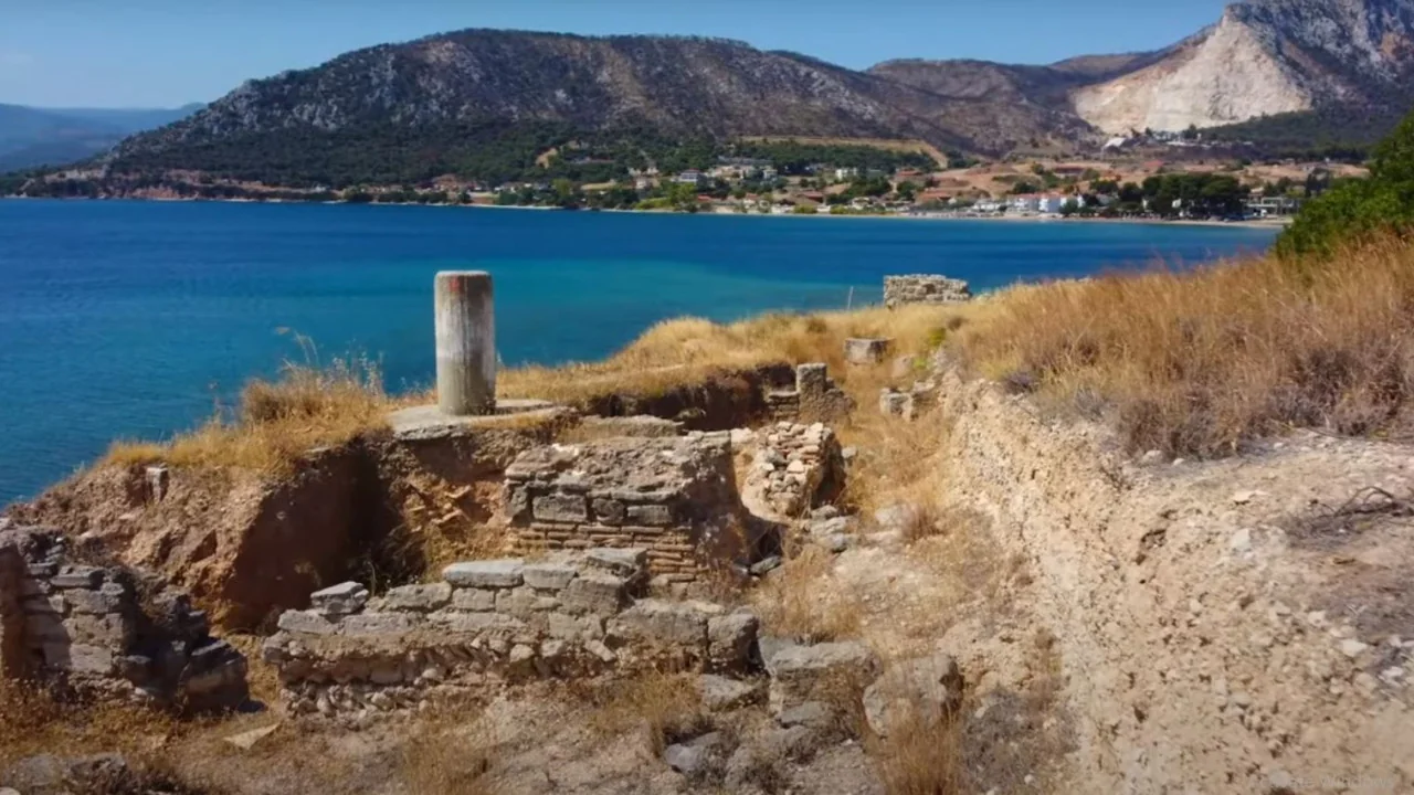 Αρχαία ερείπια και μια κολόνα πάνω από γαλάζια θάλασσα, με βουνά και ένα χωριό στο βάθος