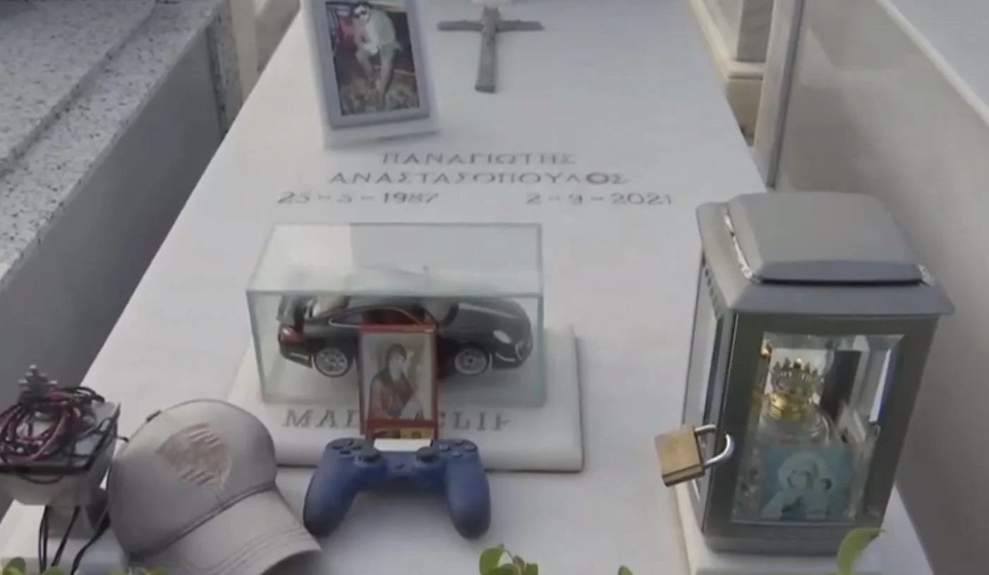 Ο λευκός μαρμάρινος τάφος του Mad Clip, με φωτογραφία, σταυρό, καντήλι, καπέλο, χειριστήριο και μινιατούρα αυτοκινήτου.