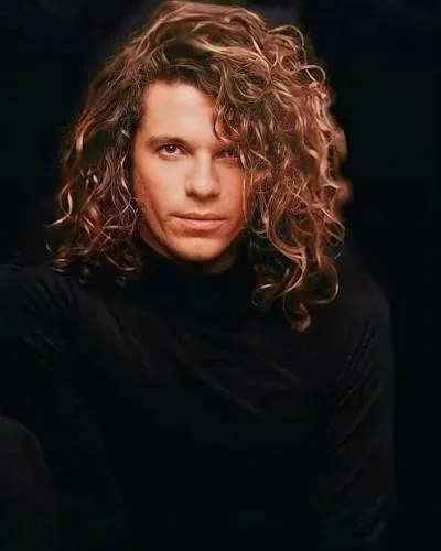 Πορτρέτο του Michael Hutchence με σγουρά μαλλιά και μαύρο πουλόβερ, σε σκούρο φόντο