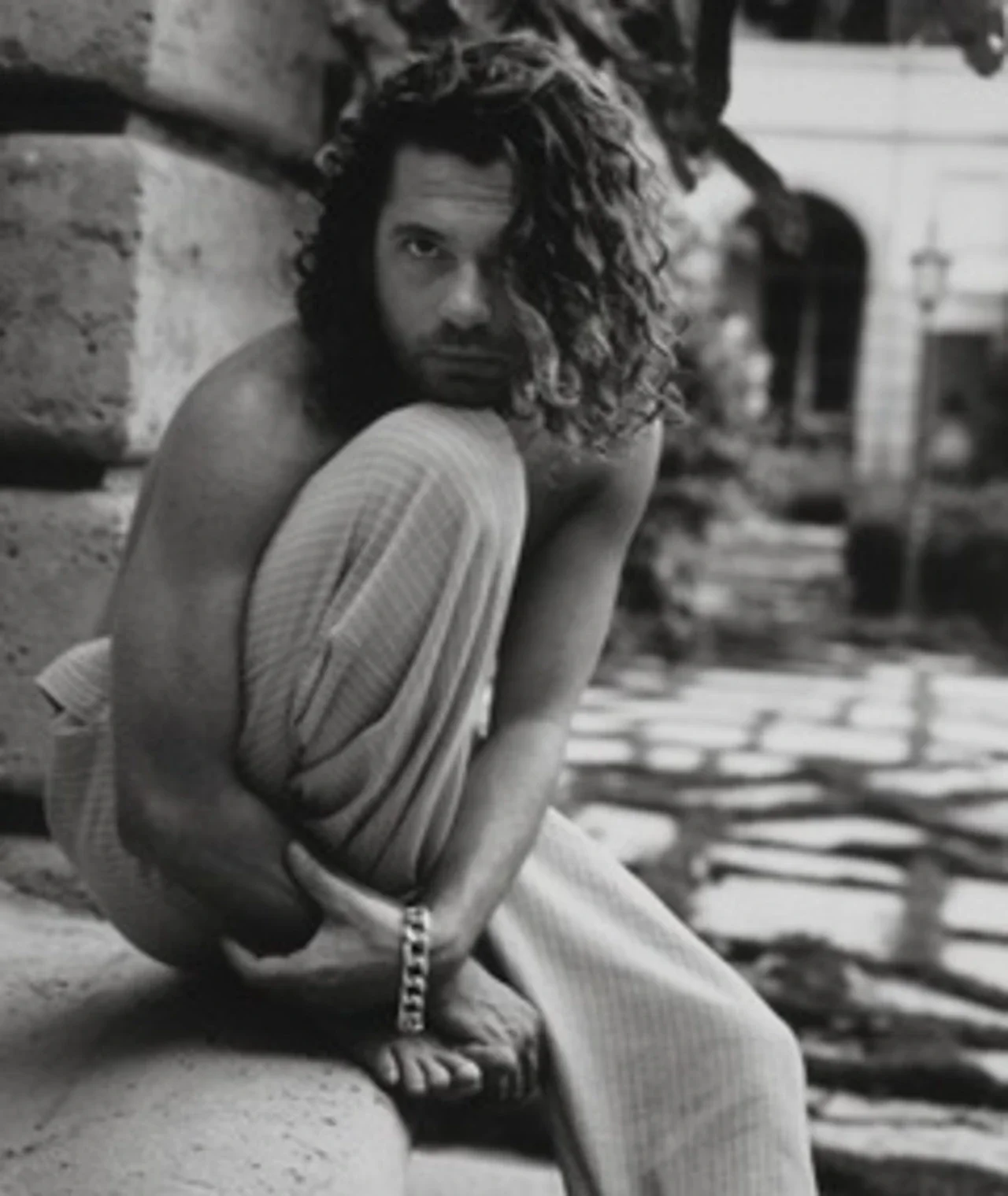 Ασπρόμαυρο πορτρέτο του Michael Hutchence, καθισμένου σε πέτρινη επιφάνεια με το χέρι γύρω από το γόνατο και βραχιόλι στον καρπό