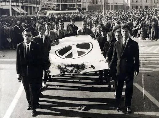 Άνδρες και γυναίκες κρατούν πανό με το σύμβολο της ειρήνης και λουλούδια, μπροστά από πυκνό πλήθος