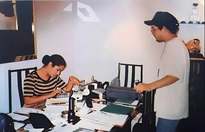 Η Selena κάθεται σε γραφείο γεμάτο χαρτιά και δουλεύει με μια συσκευή, ενώ δίπλα της στέκεται άνδρας με καπέλο