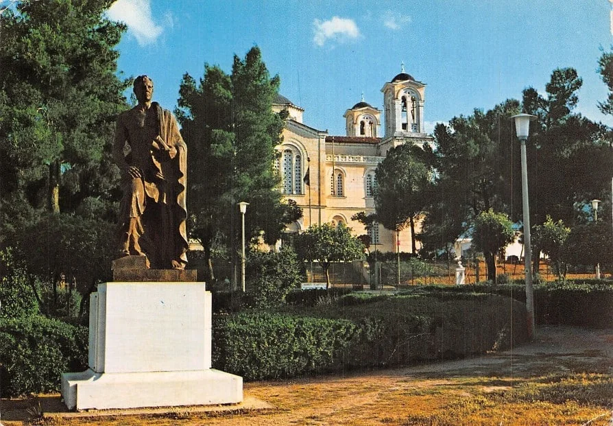 Χάλκινο άγαλμα άνδρα με μανδύα σε πάρκο, με εκκλησία με καμπαναριό πίσω του