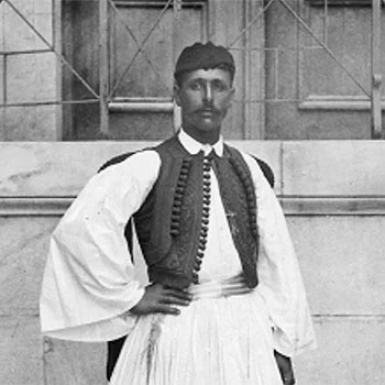 Ο Σπύρος Λούης με φουστανέλα, γιλέκο και φέσι μπροστά από παράθυρο.