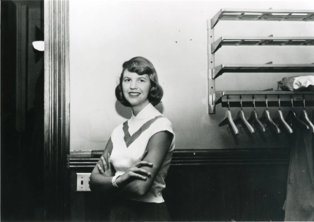Η Sylvia Plath χαμογελά με σταυρωμένα χέρια σε εσωτερικό χώρο.