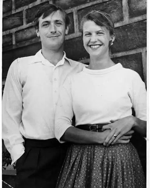 Η Sylvia Plath και ο Ted Hughes χαμογελούν, όρθιοι μπροστά σε πέτρινο τοίχο.