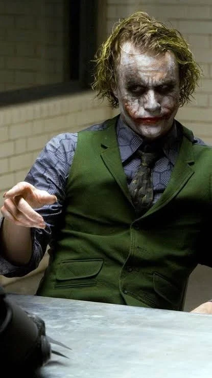 Ο χαρακτήρας του Joker με πράσινο γιλέκο και βαμμένο πρόσωπο, καθισμένος σε τραπέζι ανακριτηρίου, σε κάδρο από την ταινία «The Dark Knight»
