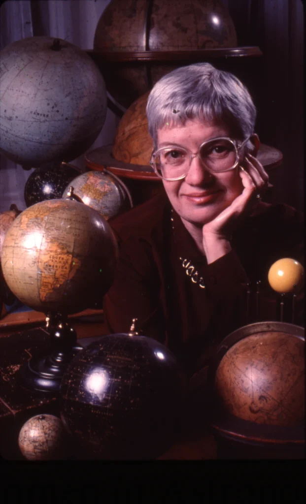 Η Vera Rubin καθισμένη ανάμεσα σε παλιές υδρόγειες σφαίρες, με το χέρι στο πρόσωπό της.