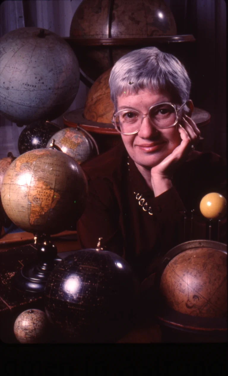 Η Vera Rubin καθισμένη ανάμεσα σε παλιές υδρόγειες σφαίρες, με το χέρι στο πρόσωπό της.