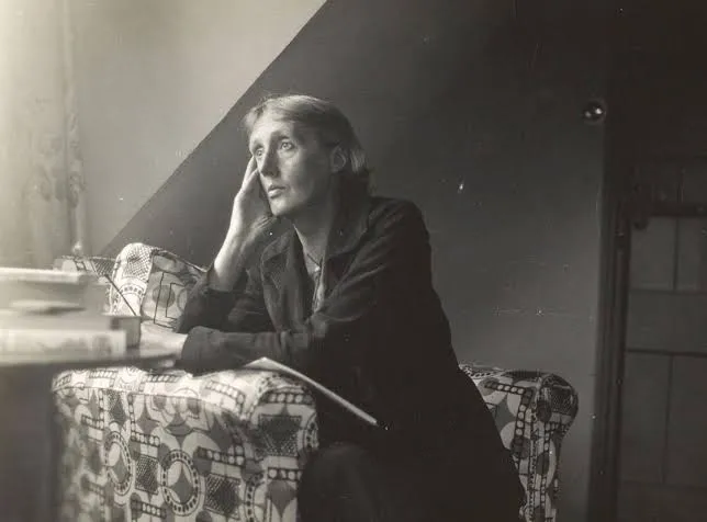 Η Virginia Woolf καθισμένη σε τραπέζι δίπλα σε παράθυρο, με το χέρι στο κεφάλι και το βλέμμα προς τα πάνω