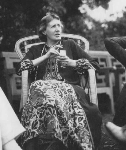 Η Virginia Woolf καθισμένη σε ψάθινη πολυθρόνα, με μοτίβο στη φούστα και ένα φλιτζάνι στα χέρια