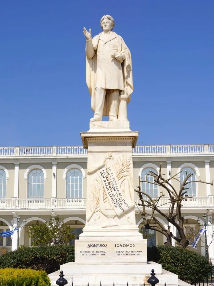 Το μαρμάρινο άγαλμα του Διονυσίου Σολωμού στη Ζάκυνθο, με υψωμένο το ένα χέρι, μπροστά από λευκό κτίριο και γαλάζιο ουρανό