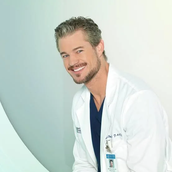 Ο Eric Dane ενσαρκώνει τον Δρ. Mark Sloan. Φοράει λευκή ιατρική ρούμπα πάνω από σκούρα μπλε scrubs και έχει μια καρτελίτσα με το όνομά του. Χαμογελάει πλατιά κοιτάζοντας την κάμερα.