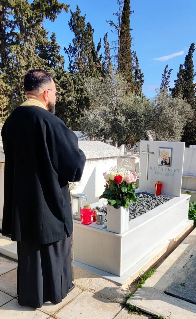 Λευκός μαρμάρινος τάφος της Καίτης Γκρέυ με σταυρό, φωτογραφία, τριαντάφυλλα και κανδήλια, και ιερέας μπροστά του