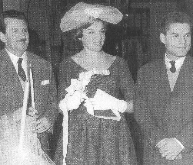 Ασπρόμαυρη φωτογραφία νεαρής γυναίκας με καπέλο με βέλο, γάντια και μπουκέτο, ανάμεσα σε δύο άνδρες, σε εκκλησία