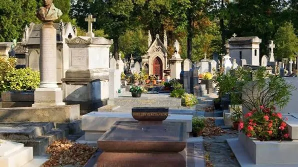 Τάφοι με σταυρούς και λουλούδια κάτω από τα δέντρα του νεκροταφείου Μονπαρνάς, με προτομή σε στήλη αριστερά