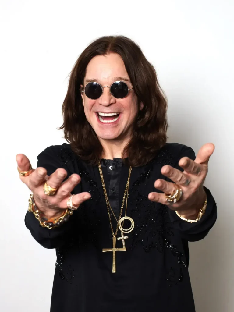 Έγχρωμο πορτρέτο του Ozzy Osbourne που χαμογελά πλατιά με ανοιχτά τα χέρια, φορώντας στρογγυλά γυαλιά ηλίου και σταυρούς