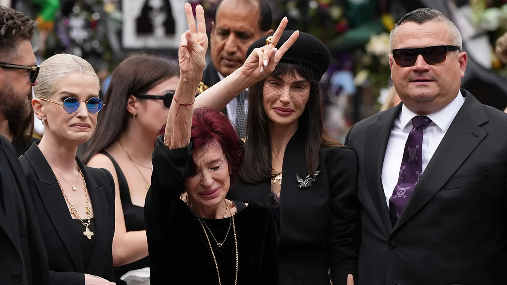 Η Sharon Osbourne κλαίει υψώνοντας τα δάχτυλα σε σχήμα V για την ειρήνη, περιστοιχισμένη από τα παιδιά της
