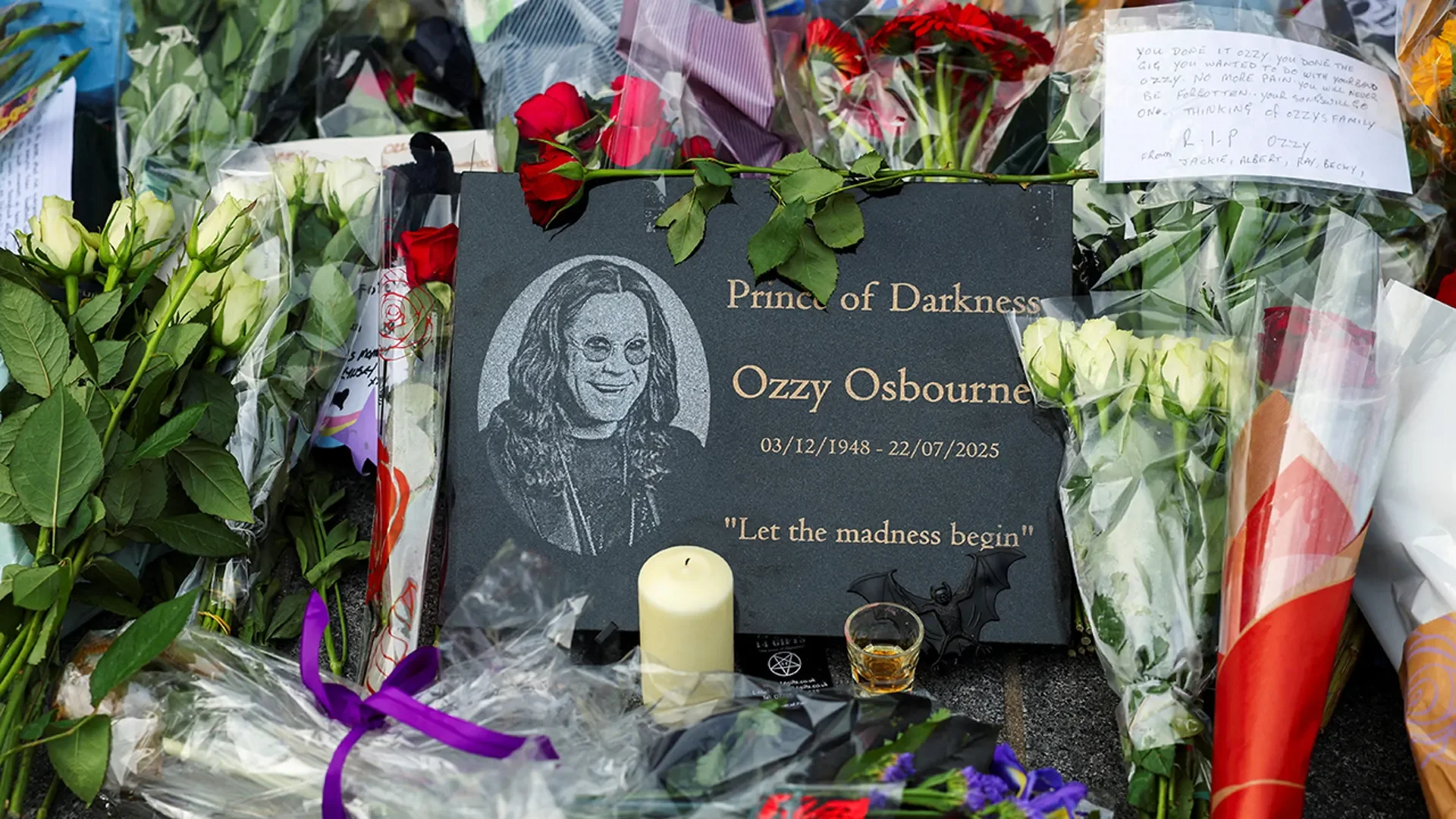 Αναμνηστική μαύρη πλάκα με το πρόσωπο του Ozzy, τις ημερομηνίες του και τη φράση Let the madness begin, ανάμεσα σε λουλούδια