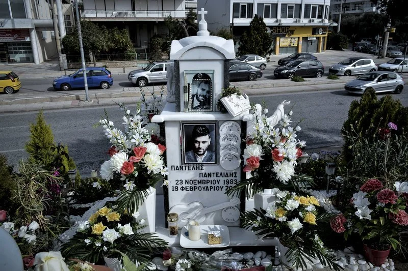 Λευκό μαρμάρινο μνημείο με σταυρό, φωτογραφίες του τραγουδιστή και στεφάνια από λουλούδια στην άκρη του δρόμου, με το όνομα Παντελής Παντελίδης και ημερομηνίες