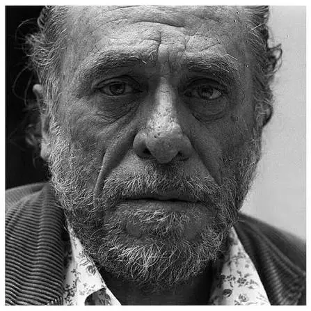 Κοντινό ασπρόμαυρο πορτρέτο του Τσαρλς Μπουκόφσκι σε μεγάλη ηλικία, με γένια, χαρακτηριστικά σημάδια στο πρόσωπο και πουκάμισο με λουλουδάτο γιακά